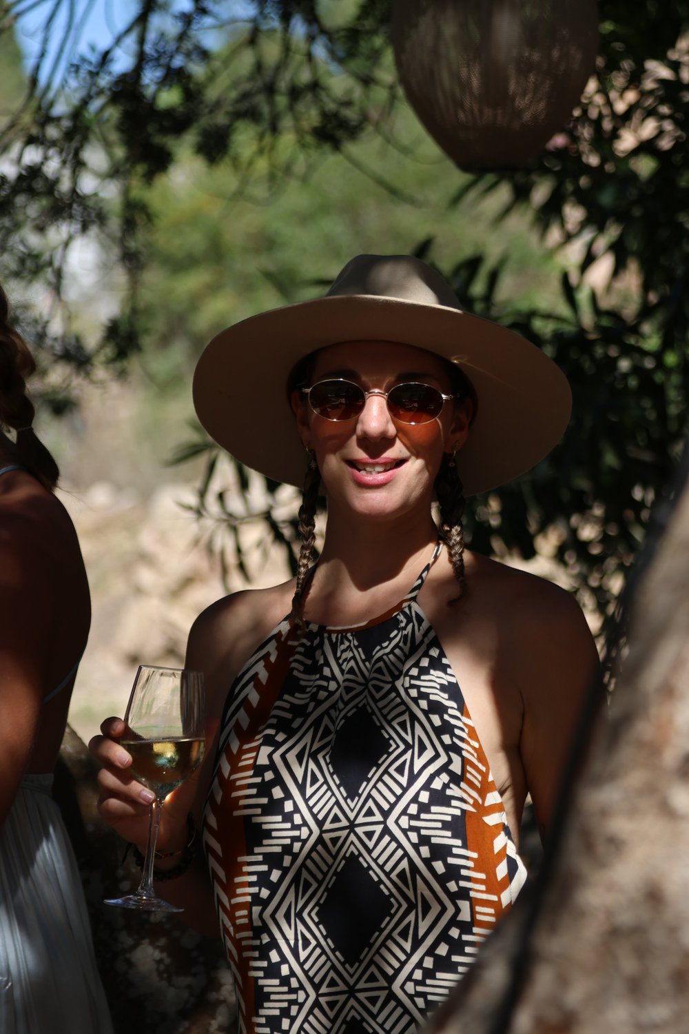 A guest in a wide sunhat holds a glass of white wine among the trees.