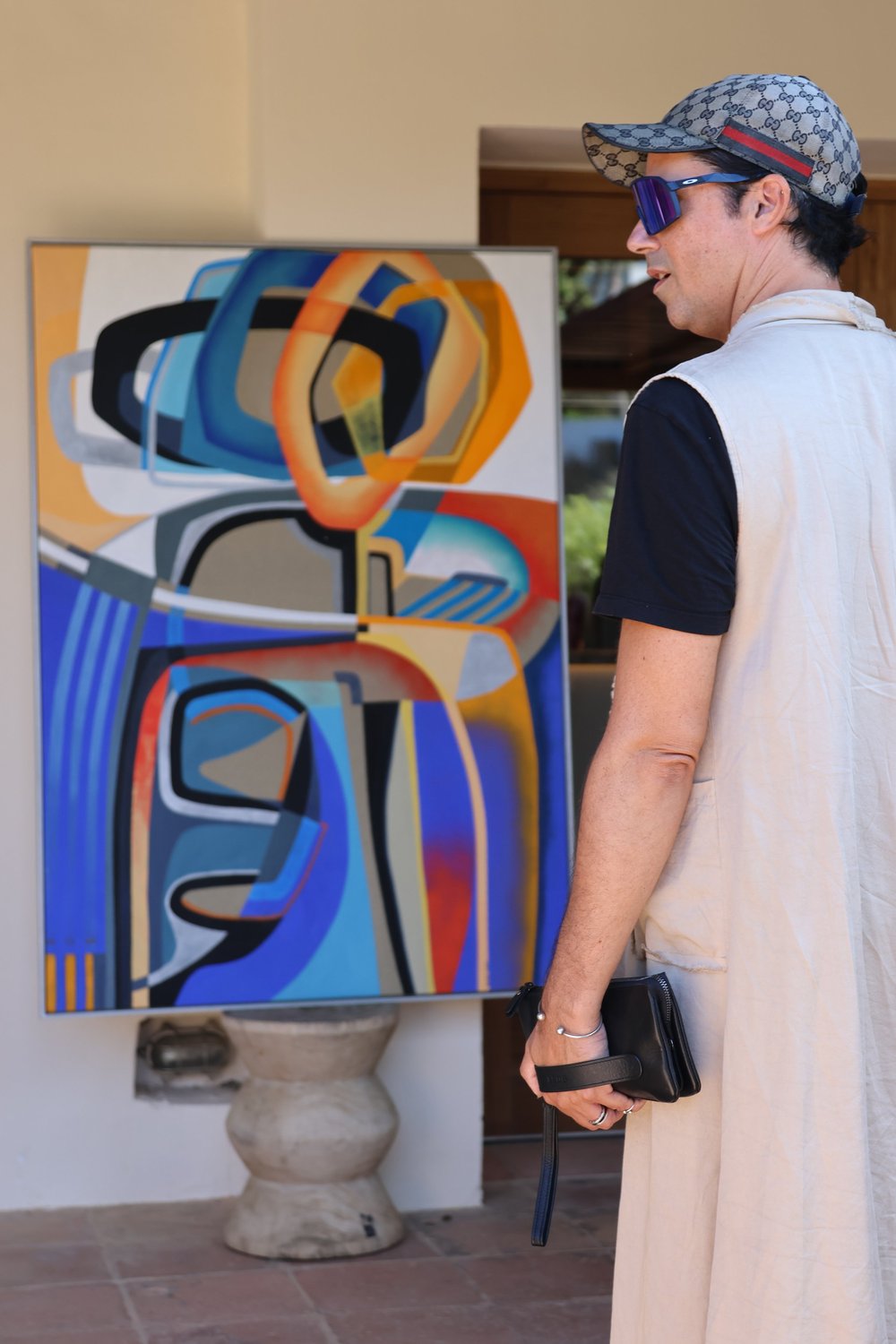 A guest in sunglasses and a cap studies an abstract painting outdoors.