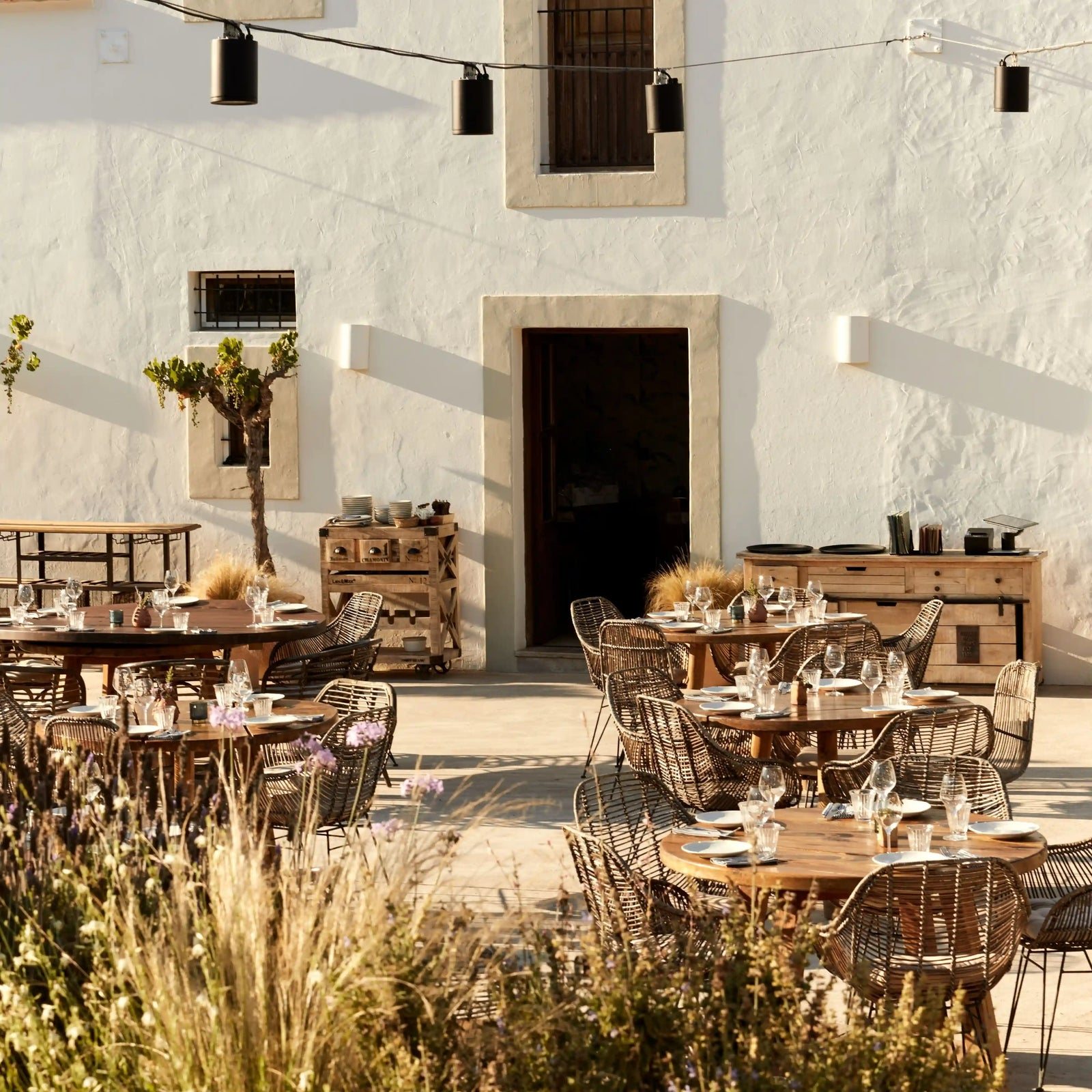 The courtyard terrace of Juntos House in Sant Mateu d&rsquo;Albarca: lunch tables set before a white traditional Ibizan house, with wild grasses and string lights.