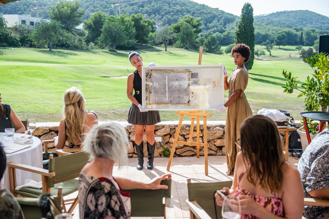 Two presenters show an artwork on an easel to guests seated on the lawn.