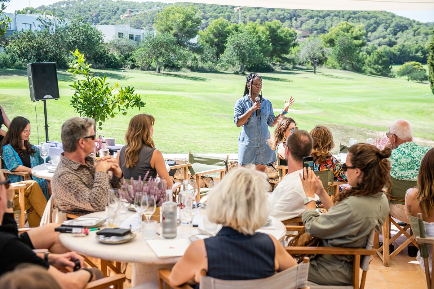 A speaker with a microphone addresses guests on a lawn overlooking the hills.